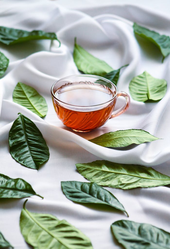 A clear glass cup of tea surrounded by green leaves on a white silky fabric, creating a calming scene.