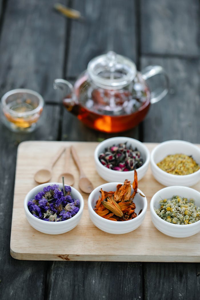 A variety of dried herbs and flowers in bowls with a glass teapot on a wooden board.